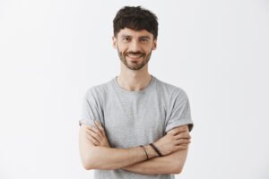 Waist-up shot of confident good-looking mature european male entrepreneur in t-shirt holding hands crossed in self-assured pose and smiling with assured pleasant smile over gray background